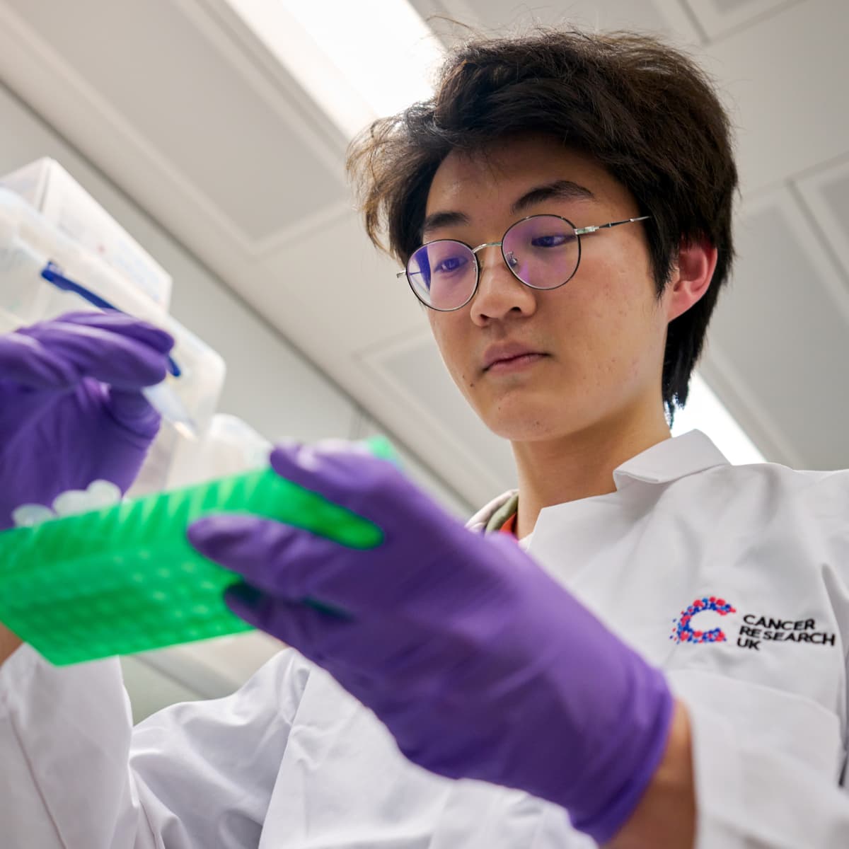A scientist in the lab looking down at some test tubes. They're wearing glasses, a lab coat and purple gloves.