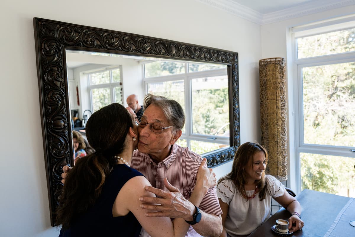 A photo of an older person with glasses that embraces and kisses a younger person on the cheek in a bright living room.