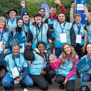 Large group of about 20 volunteers huddled together and smiling to the camera. Gathered by the finish line at the end of an event course.